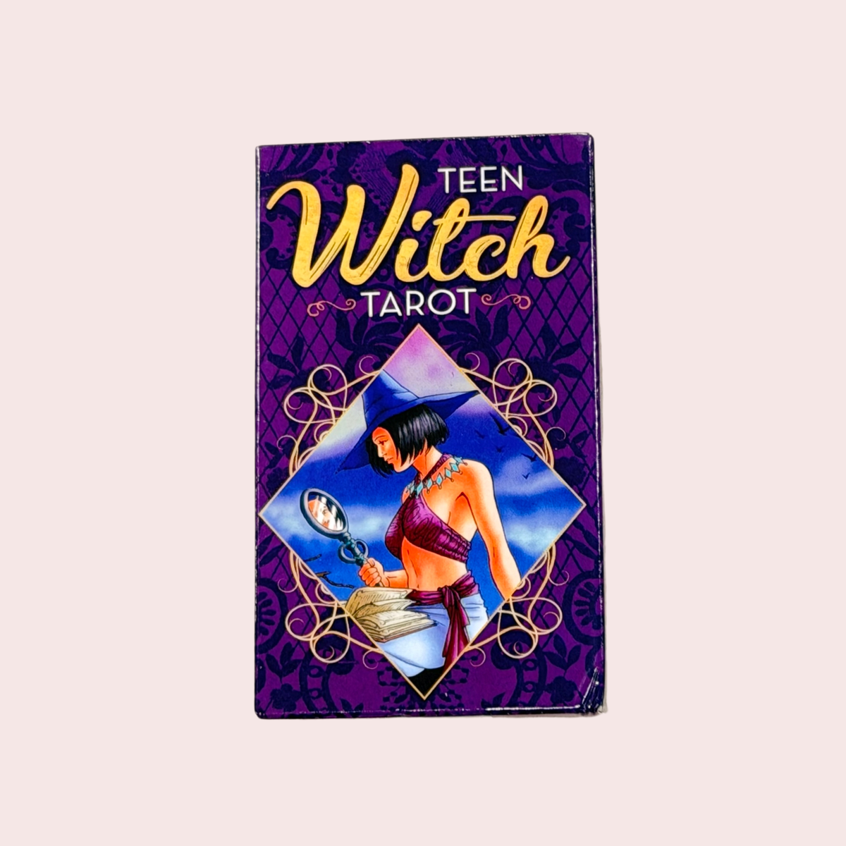 Caja del Teen Witch Tarot, morada con detalles dorados, sobre fondo rosa